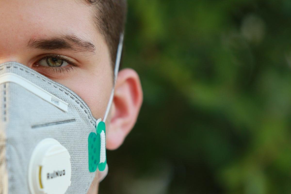 Un jeune homme pose avec un masque sanitaire sur le visage, nous ne voyons que la moitié gauche de son visage ; un droite un fond flou de verdure.