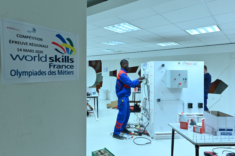Au premier plan à gauche, l'affiche des World Skills 2020 épreuve régionale d'électricité, au second plan, un jeune au travail sur une maquette.