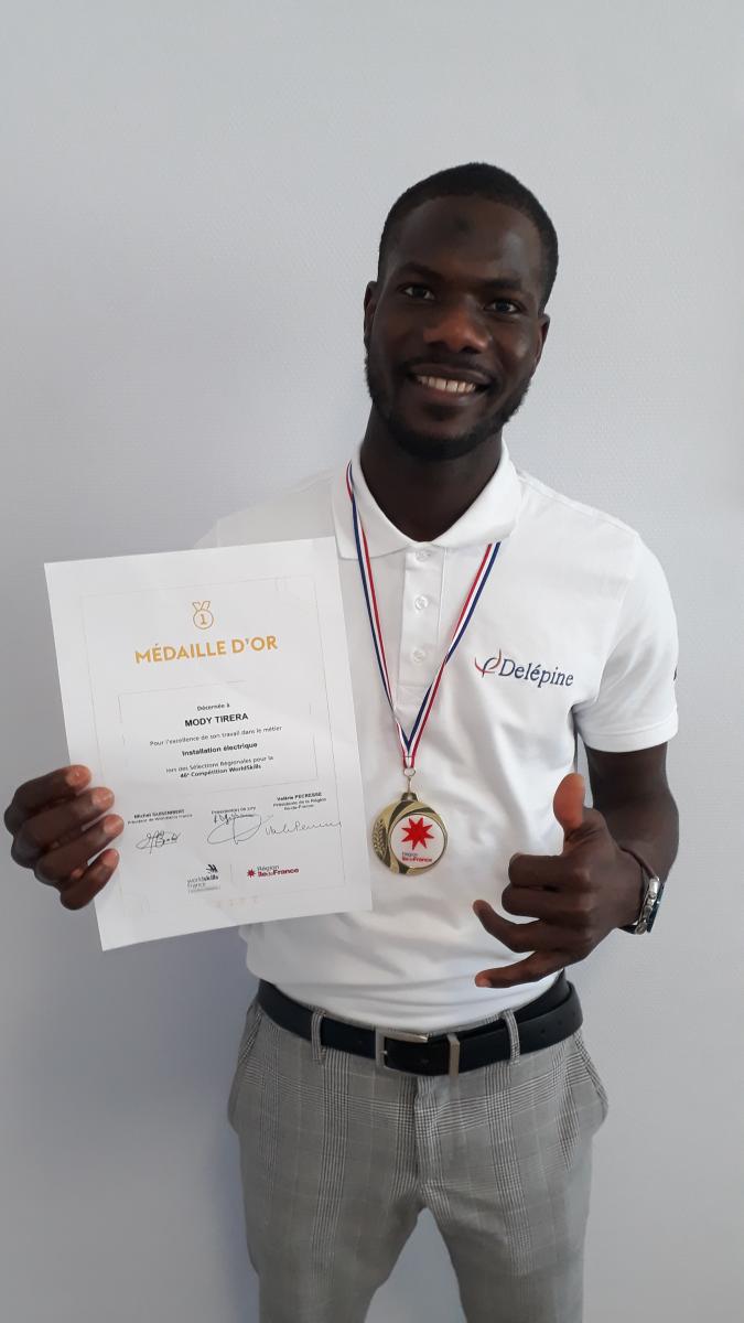 Mody la médaille d'or des World Skills pose fièrement avec son diplôme, tout sourire, un pouce en l'air.